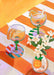 Two margarita glasses with orange slices and straws rest on a striped table beside a vase of white flowers, styled next to the Hôtel Dodo "A L'Orange Giclée Art Print.