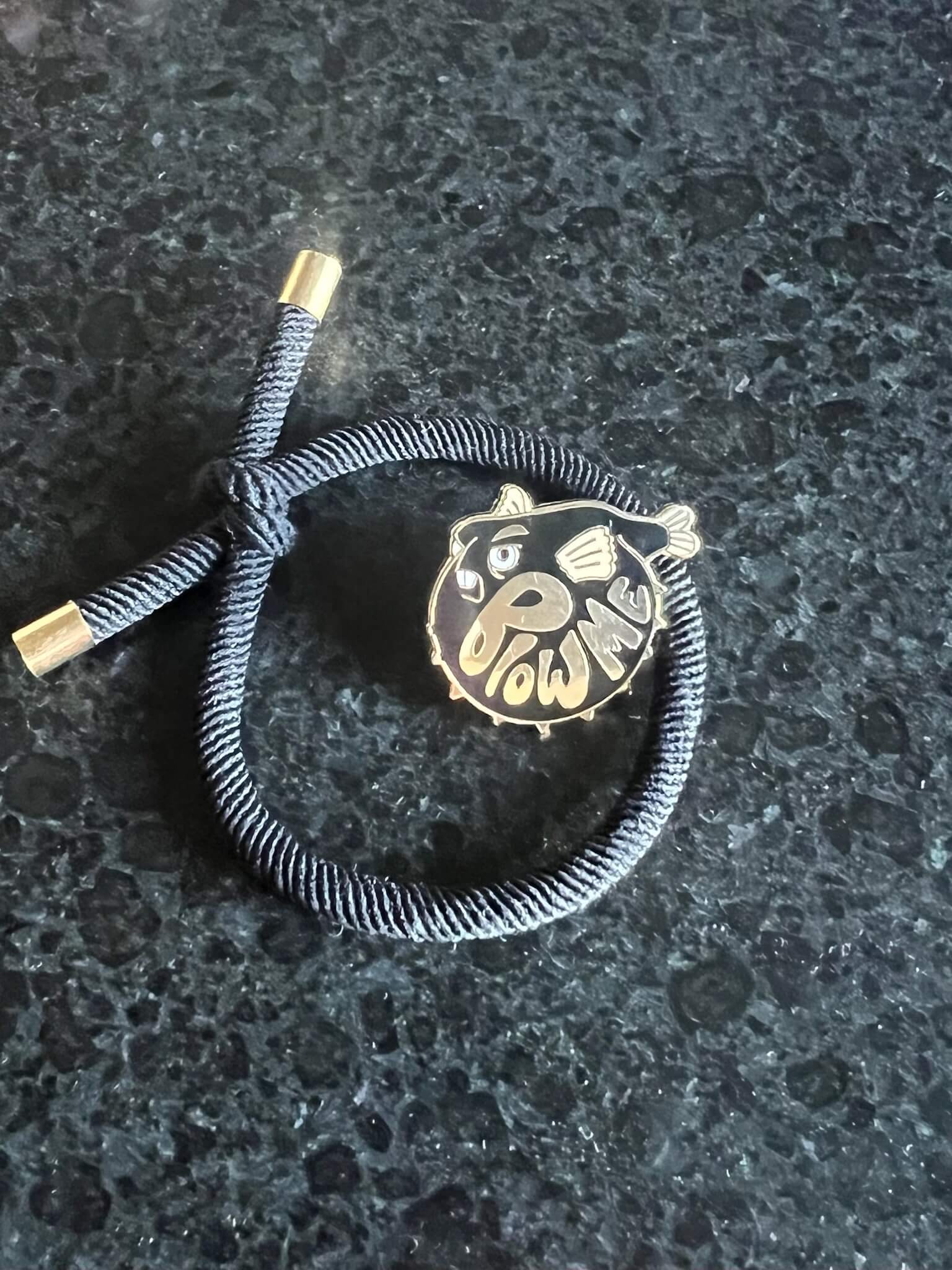 A dark bracelet sits on a deep surface, featuring a gold-accented knot and a round charm with the Pins by diedododa Beastly Blowfish cartoon, alongside the playful text "WOW ME.