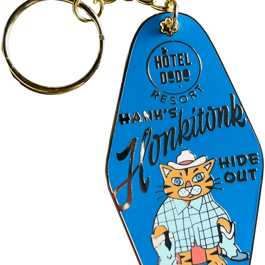 The Hank's Honkitonk Hideout Keyring, from Hôtel Dodo, is a blue keychain featuring a cat in a cowboy hat and shirt, labeled "Hank's Honkitonk," with an enamel tag reading "Hide Out," capturing the charm of a rustic log cabin retreat.