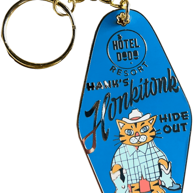 The Hank's Honkitonk Hideout Keyring, from Hôtel Dodo, is a blue keychain featuring a cat in a cowboy hat and shirt, labeled "Hank's Honkitonk," with an enamel tag reading "Hide Out," capturing the charm of a rustic log cabin retreat.