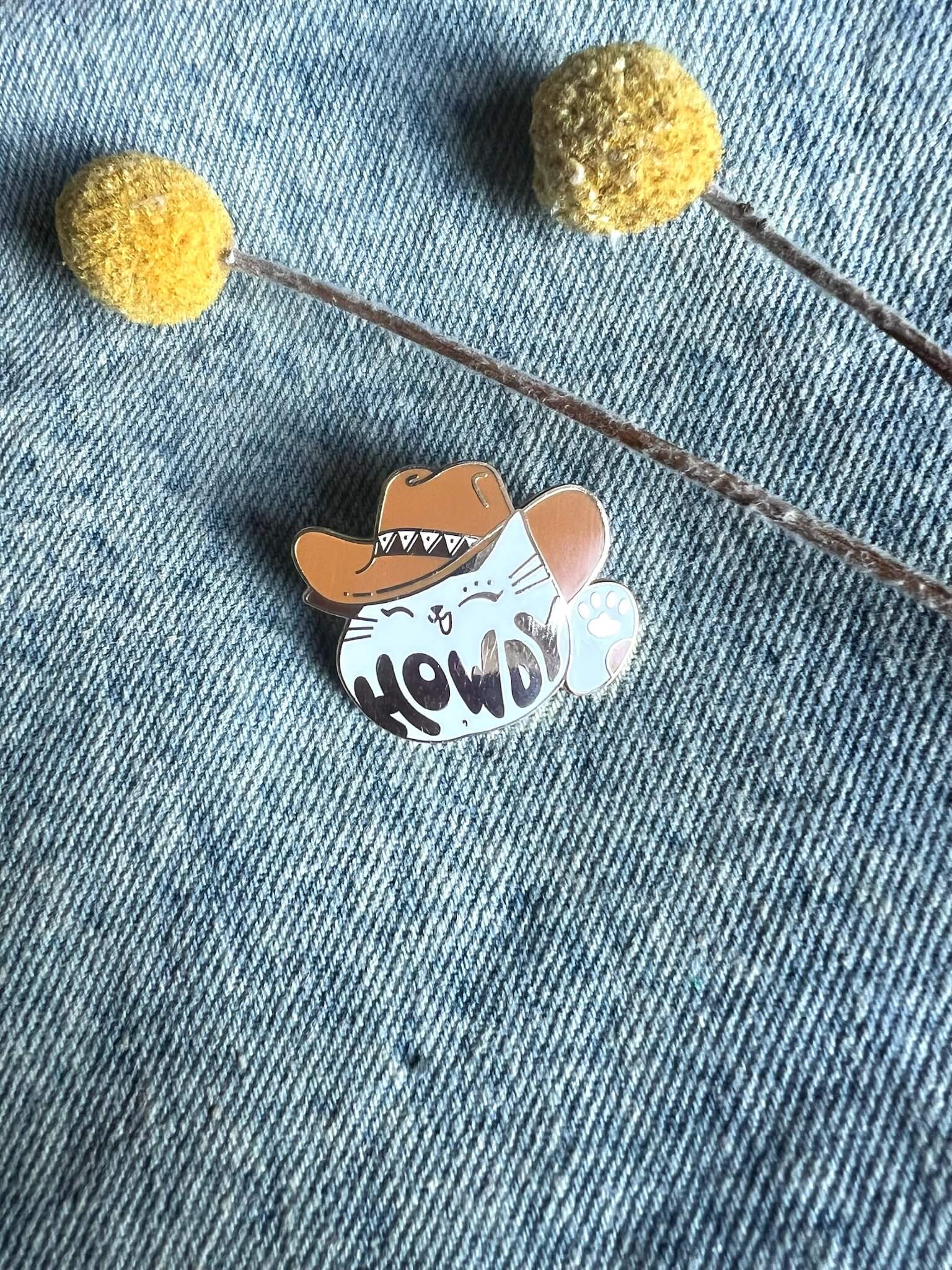 The Howdy Pawdner pin by Pins by diedododa features a cartoon cat, resembling a lucky cat, wearing a cowboy hat with "HOWDY" on its belly. It rests on denim fabric, accompanied by two yellow billy ball flowers for an added touch of whimsy and good fortune.