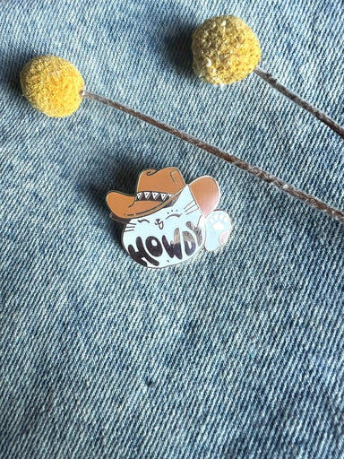 The Howdy Pawdner pin by Pins by diedododa features a cartoon cat, resembling a lucky cat, wearing a cowboy hat with "HOWDY" on its belly. It rests on denim fabric, accompanied by two yellow billy ball flowers for an added touch of whimsy and good fortune.