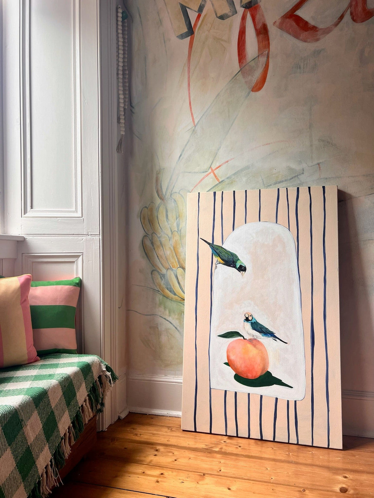 The Peach Treaty Giclée Canvas Print by Avian Alliances features two colorful birds and a peach, displayed against an abstract mural beside a window seat with striped cushions and a green checkered blanket.