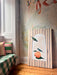 The Peach Treaty Giclée Canvas Print by Avian Alliances features two colorful birds and a peach, displayed against an abstract mural beside a window seat with striped cushions and a green checkered blanket.