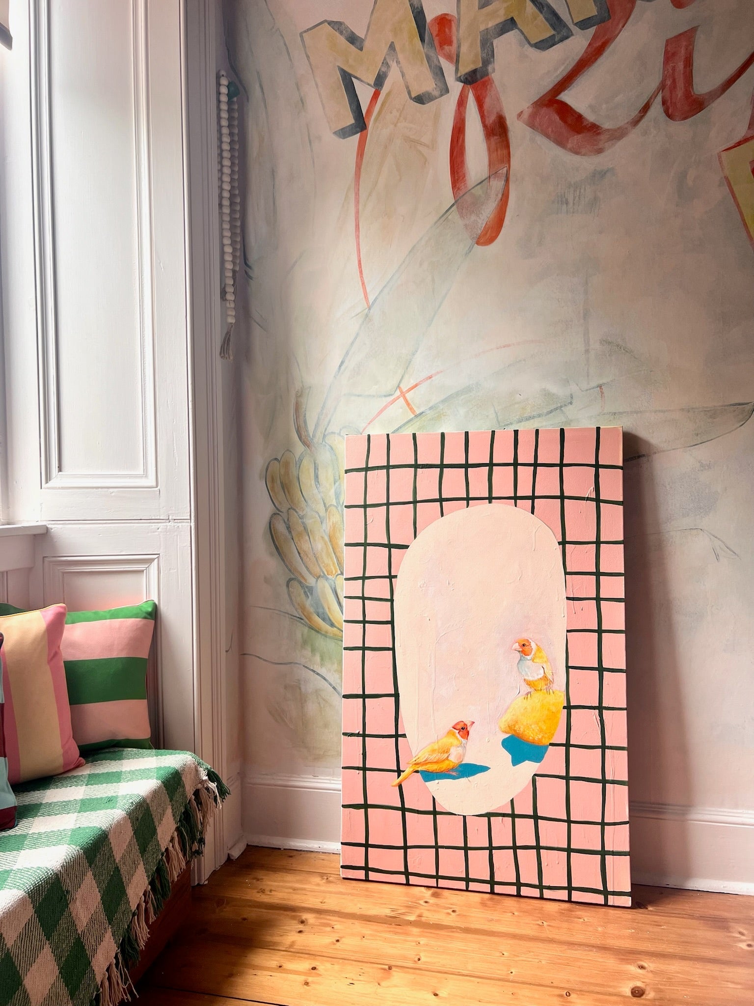 The Bittersweet Conferrals Giclée Canvas Print by Avian Alliances features two birds on a pink grid, adding vibrant charm to a window seat with striped cushions and a green-checked blanket beside mural art.