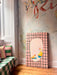 The Bittersweet Conferrals Giclée Canvas Print by Avian Alliances features two birds on a pink grid, adding vibrant charm to a window seat with striped cushions and a green-checked blanket beside mural art.