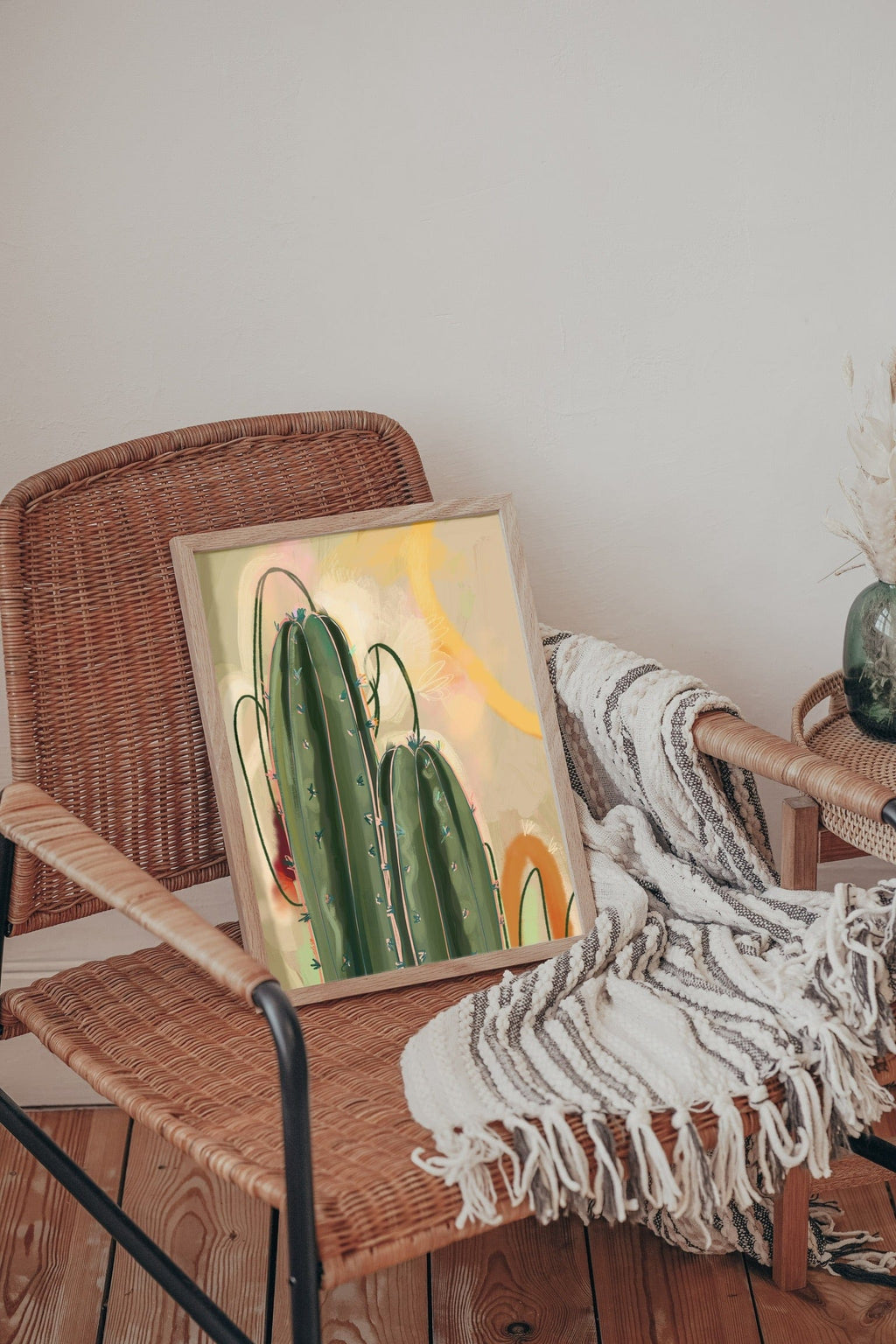 The Saguaro Sundown Giclée Art Print by Heat Flares brings desert vibes, resting on a woven chair beside a striped blanket, with a side table and dried flowers enhancing the sun-soaked scene.