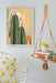 Heat Flares’ Saguaro Sundown Giclée Art Print hangs on a white wall, paired with a macramé shelf holding books and a mug. A cozy white chair accented by a yellow polka dot pillow adds comfort to the setting.