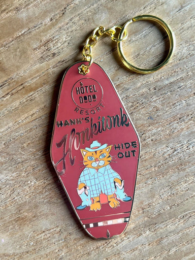 The Hank's Honkitonk Hideout Keyring by Hôtel Dodo features a vintage-style design with "Hôtel Dodo Resort" and "Hank's Honkatonk Hideout," adorned with a cartoon cowboy cat enamel tag reminiscent of a rustic log cabin getaway.