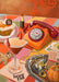 Certainly! The Harvest Club Giclée Art Print by Hôtel Dodo features an orange rotary phone, devil-horned cocktail, pumpkin, pie, lipstick, voodoo doll, tarot cards & jewelry on a colorful tiled table with autumn decor.
