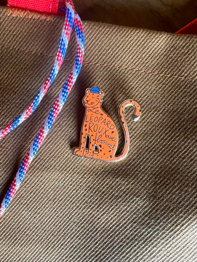 A charming ginger Léopard Roux Pin by Pins by diedododa features a freckled face and "Leopard Roux has Bonnet" on its body, attached with a stylish red and blue striped rope.