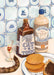 The Dutch Courage Giclée Art Print by Hôtel Dodo features a jenever bottle, a glass, "Cat Tonic," a cat tail, and a plate of waffles with chocolate sprinkles set against Delftware blue and white tiles.