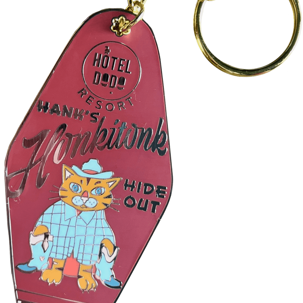 The Hank's Honkitonk Hideout Keyring by Hôtel Dodo features a vintage pink motel key tag design with a cartoon cat in a blue outfit, capturing the charm of an enamel tag reminiscent of a secluded rustic cabin.