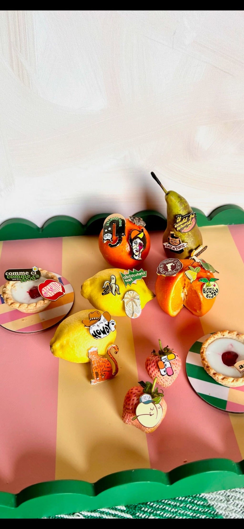 Oranges, a pear, lemon, and apple are adorned with colorful pins like the Farm Fresh Peach Pin by Pins by diedododa, displayed on a pink and green striped surface that exudes the essence of spring fashion.