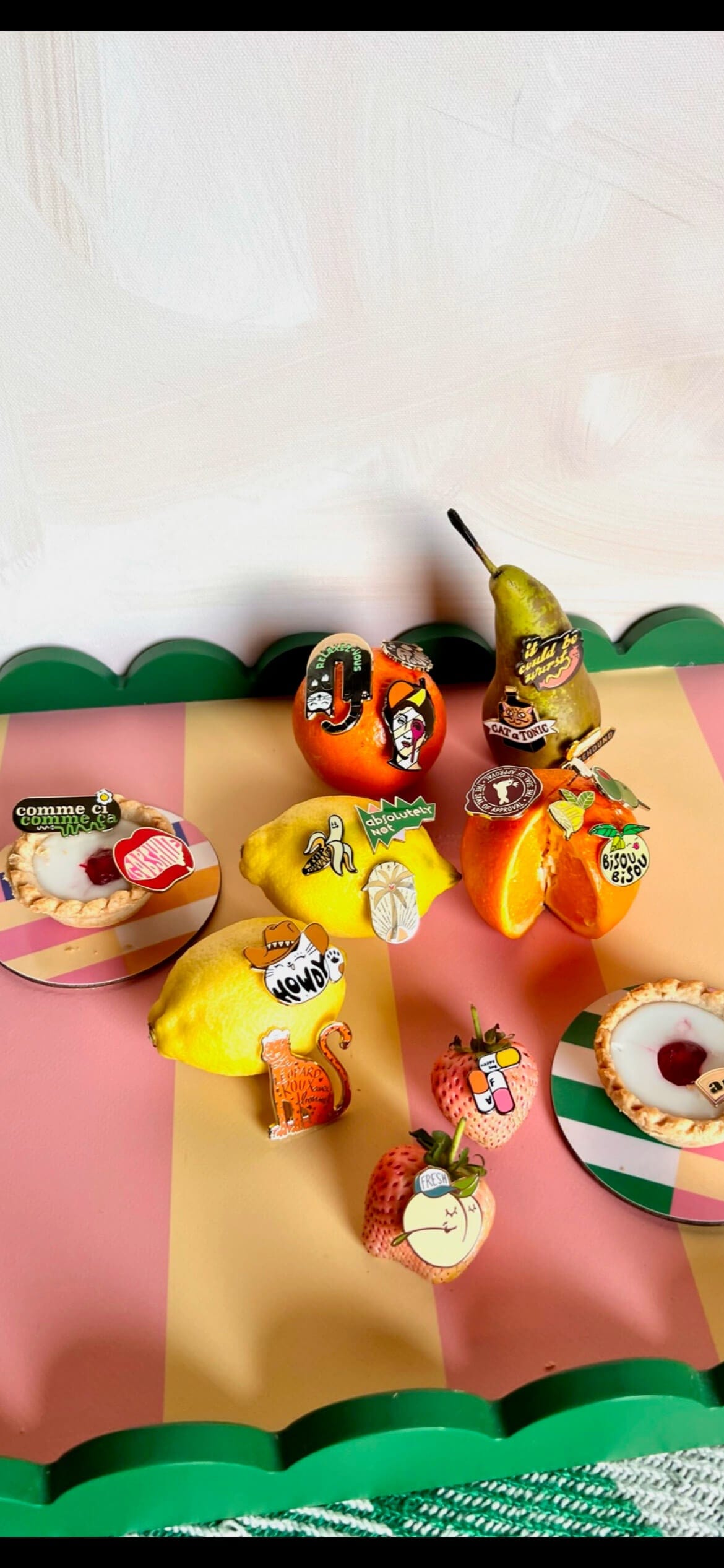 Oranges, a pear, lemon, and apple are adorned with colorful pins like the Farm Fresh Peach Pin by Pins by diedododa, displayed on a pink and green striped surface that exudes the essence of spring fashion.