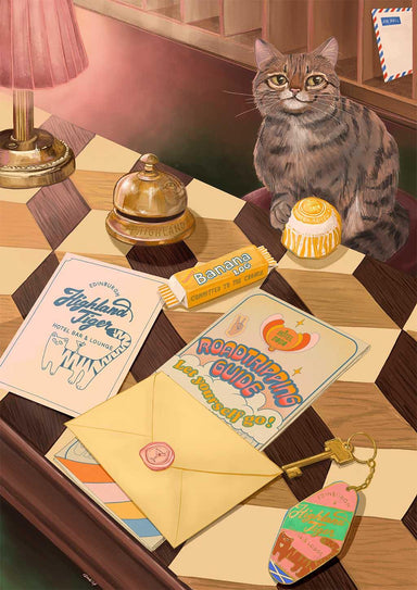 A tabby cat sits on a checkered desk surrounded by stationery, a quirky keychain, a yellow envelope, a candy bar, a cupcake, helpful travel guides titled “Road Tripping Guide” and “Edinburgh Trip,” and the Highland Tiger Lodge Giclée Art Print from Hôtel Dodo.